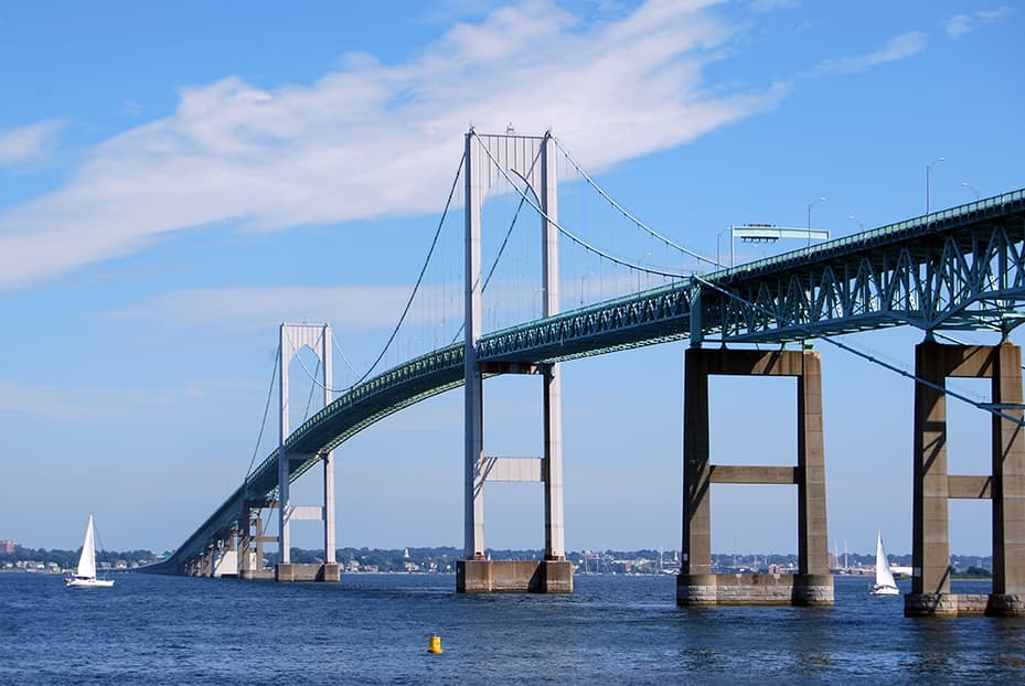 A large suspension bridge spans across a body of water with sailing cruises and sailboats below, and a city visible in the background under a blue sky.