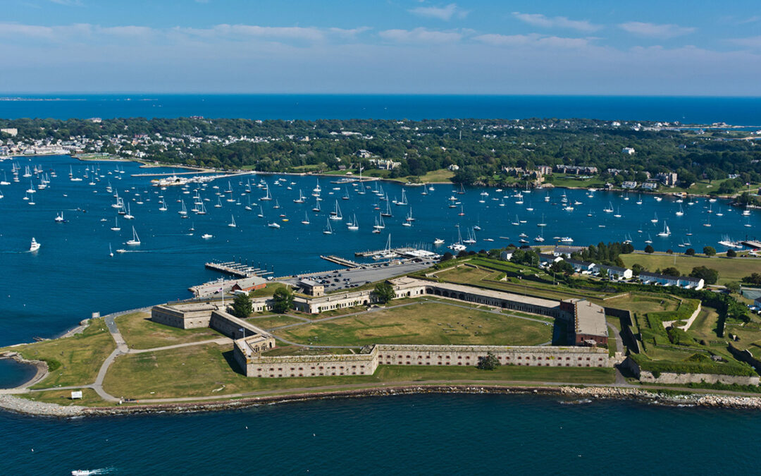 Aerial view of a historic coastal fort surrounded by water, with numerous sailboats and sailing cruises anchored in the harbor and a town visible in the background.