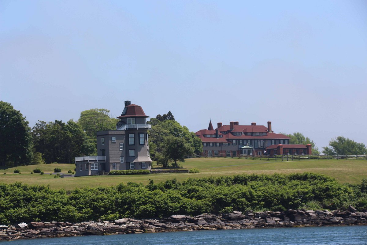 A grey lighthouse and a large red-roofed mansion sit on a grassy hill near the rocky shoreline, where sailing cruises often pass by, with trees and blue sky in the background.