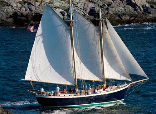 A large sailboat with two masts and white sails, perfect for sailing cruises, glides on blue water near a rocky coastline. Several people are visible on the deck.